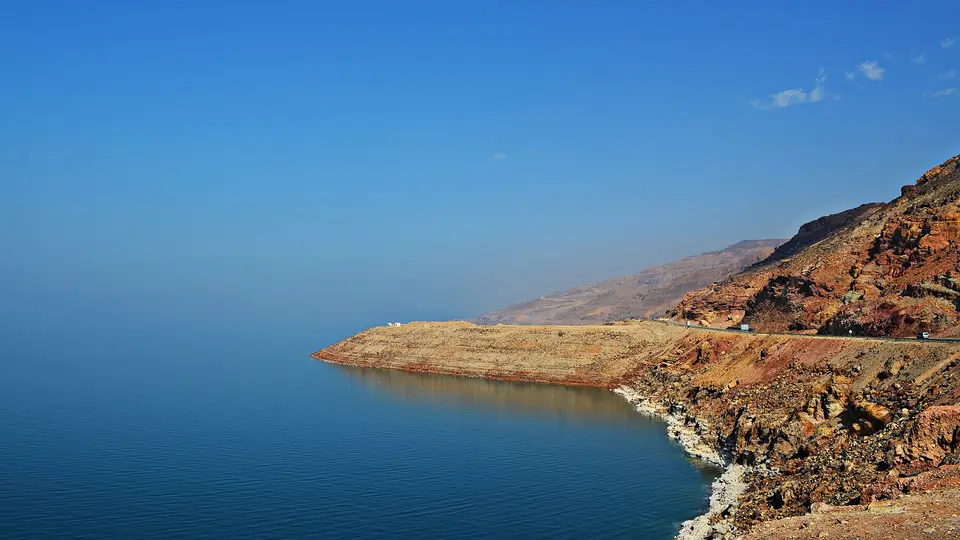 The calm blue Dead Sea below rust-coloured cliffs and the coast road on the Jordanian shore