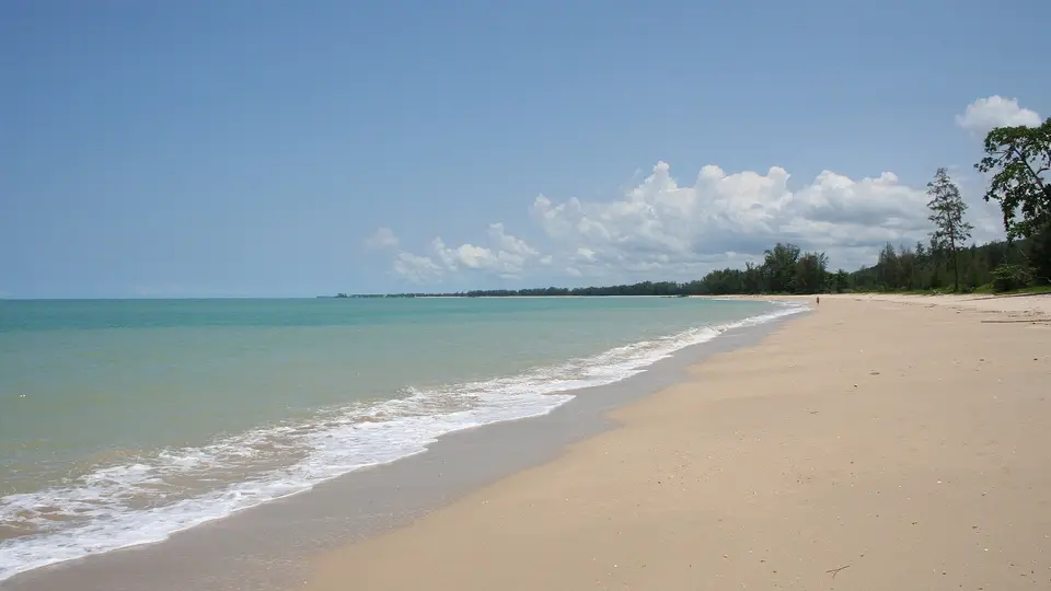 A long, empty sandy beach at Khao Lak with gentle surf and trees at the far end