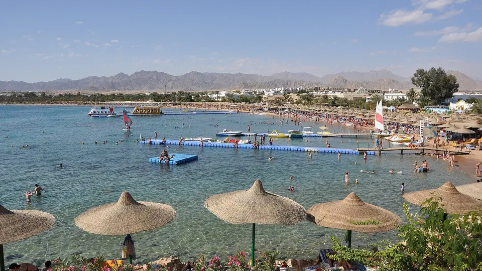 Straw parasols and swimmers along the shallow water of Naama Bay, with mountains behind