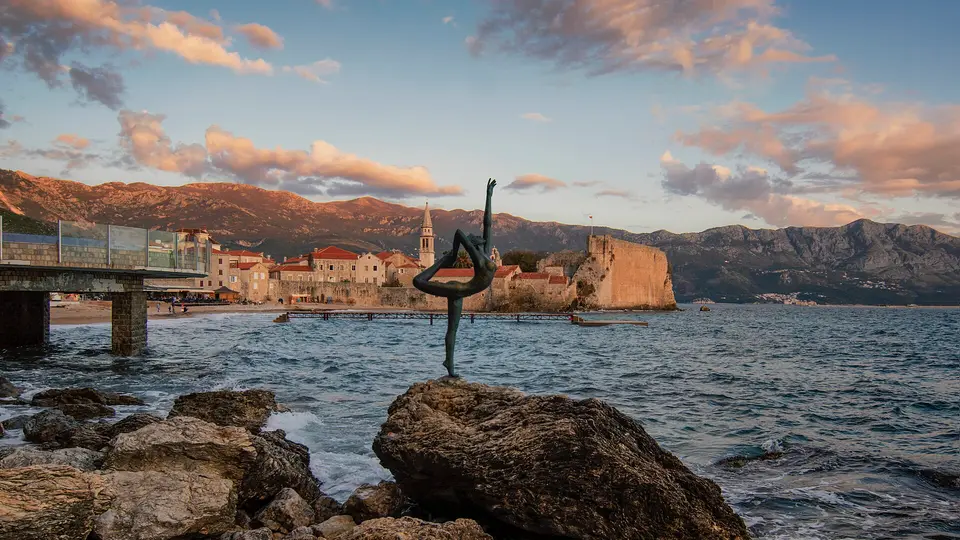 The bronze dancer statue on a seafront rock, with Budva Old Town and mountains behind at sunset